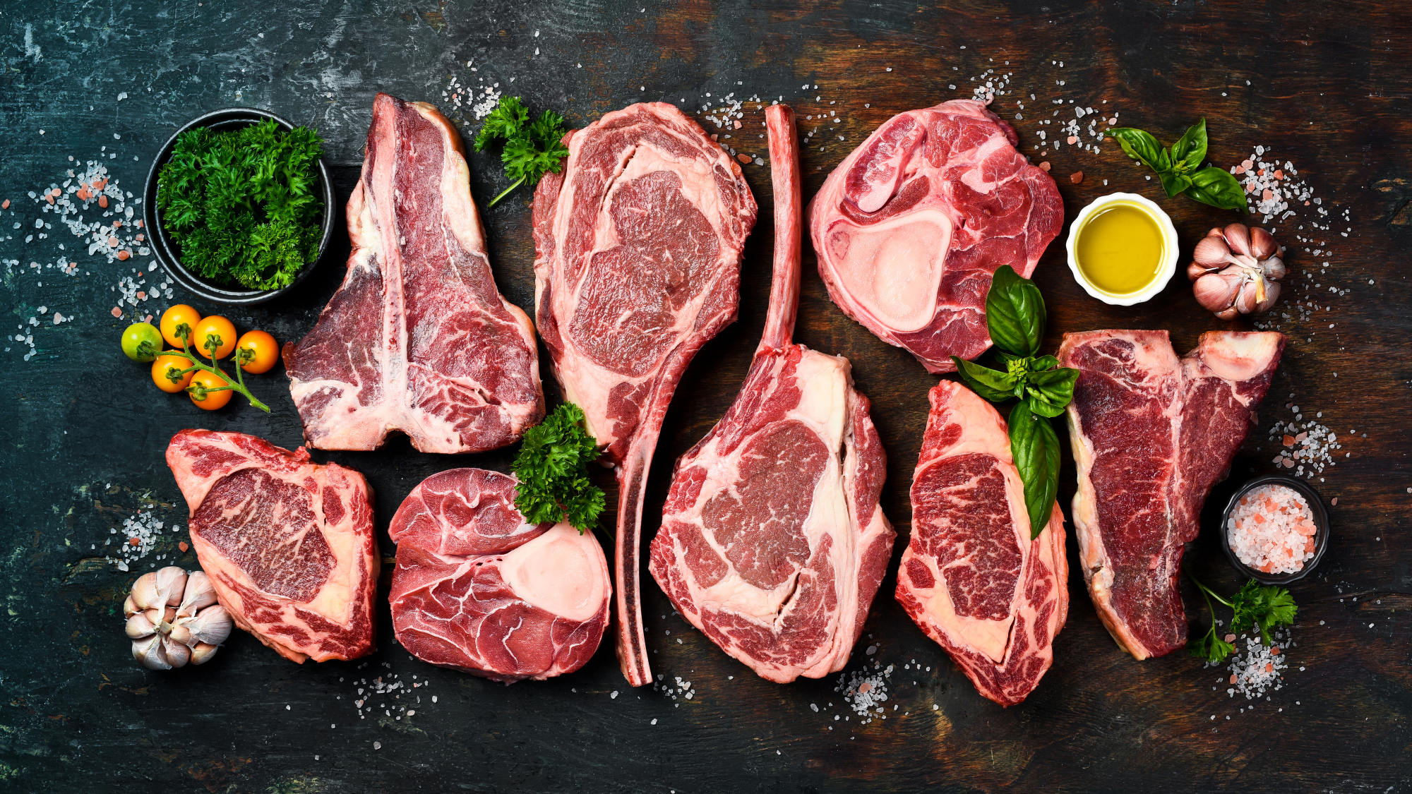 Assorted cuts of raw beef, including ribeye and tomahawk steaks, arranged on a wooden surface with fresh herbs, garlic, cherry tomatoes, olive oil, and sea salt for a gourmet cooking presentation.